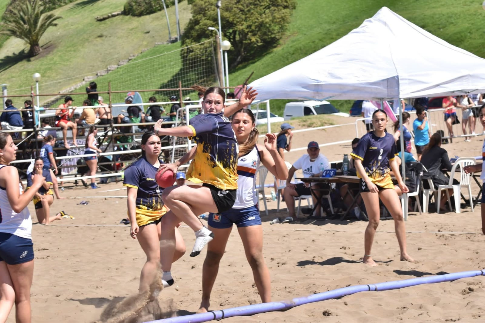 Jugadora de la Escuela Municipal de Handball de Patagones salta para lanzar al arco durante un partido de beach handball en la arena, ante la marca de una rival y con público y sombrillas de fondo