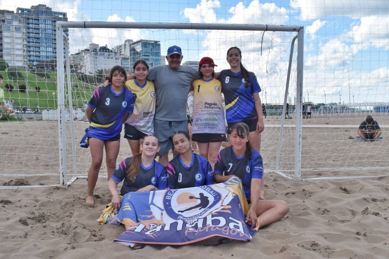 Equipo femenino de beach handball de Patagones posa delante del arco con la bandera del beach handball patagónico junto a su entrenador