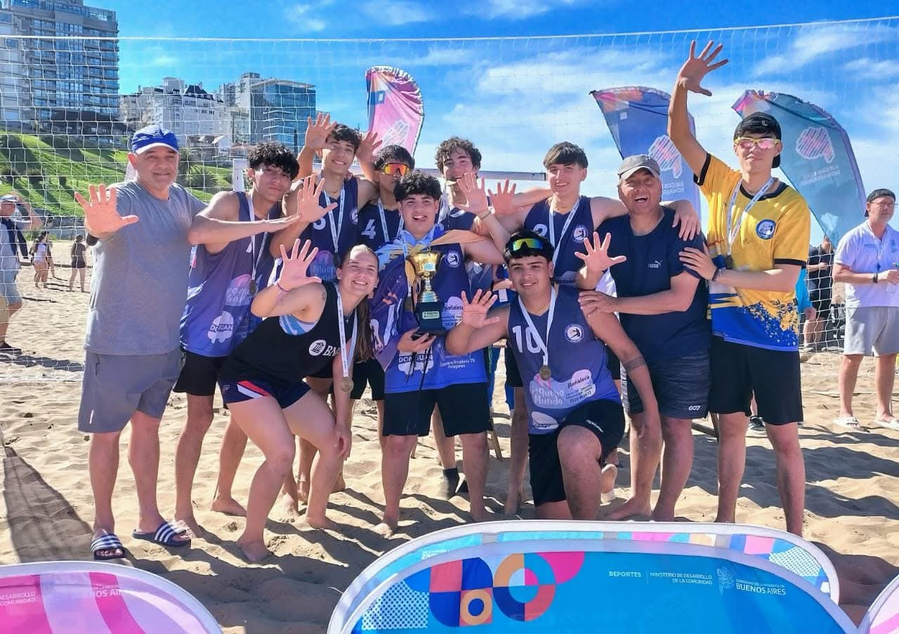 Jugadores y entrenadores de la Escuela Municipal de Handball de Patagones celebran con el trofeo y las medallas de los Juegos Bonaerenses en la arena, junto a banderas del torneo