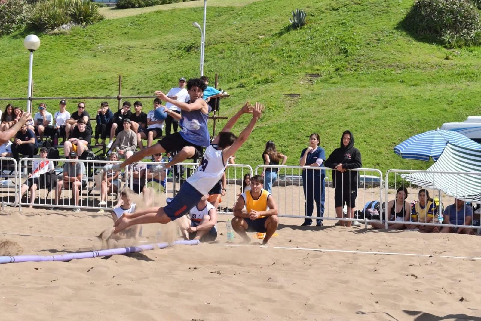 Jugador juvenil de Patagones define en el aire durante un partido de beach handball sobre la arena, con la tribuna y el público detrás de la reja