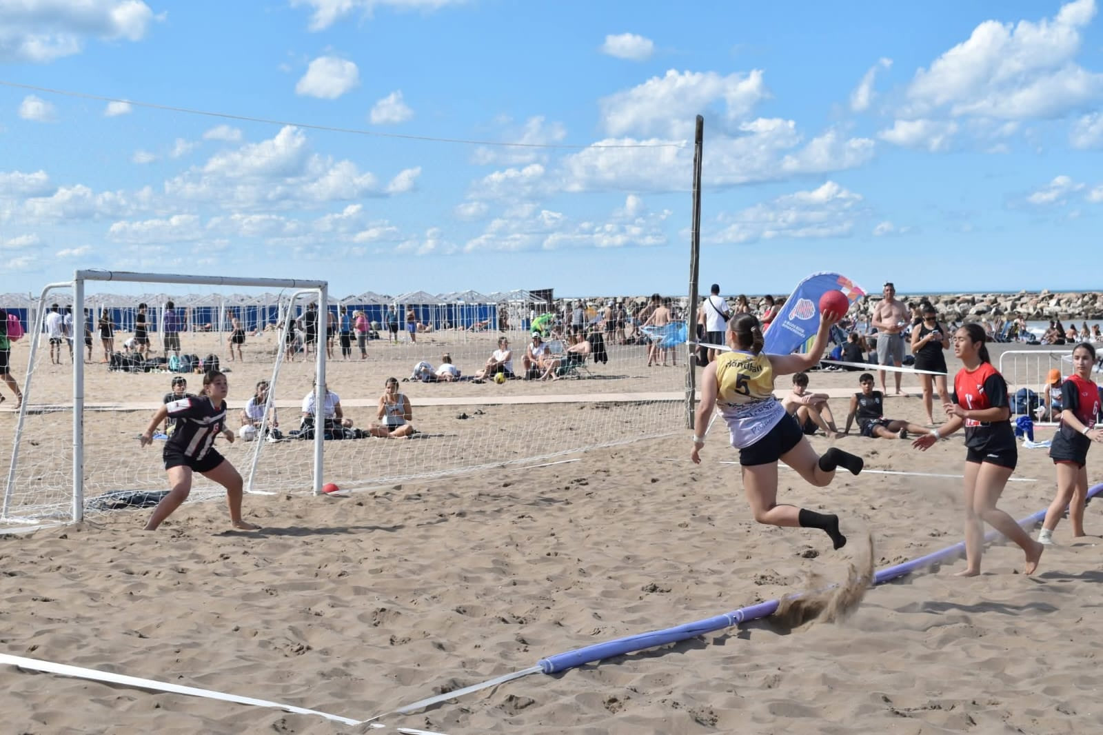 Jugadora número 5 de Patagones salta con la pelota para definir en un partido de beach handball en la playa, frente al arco y con el público alrededor