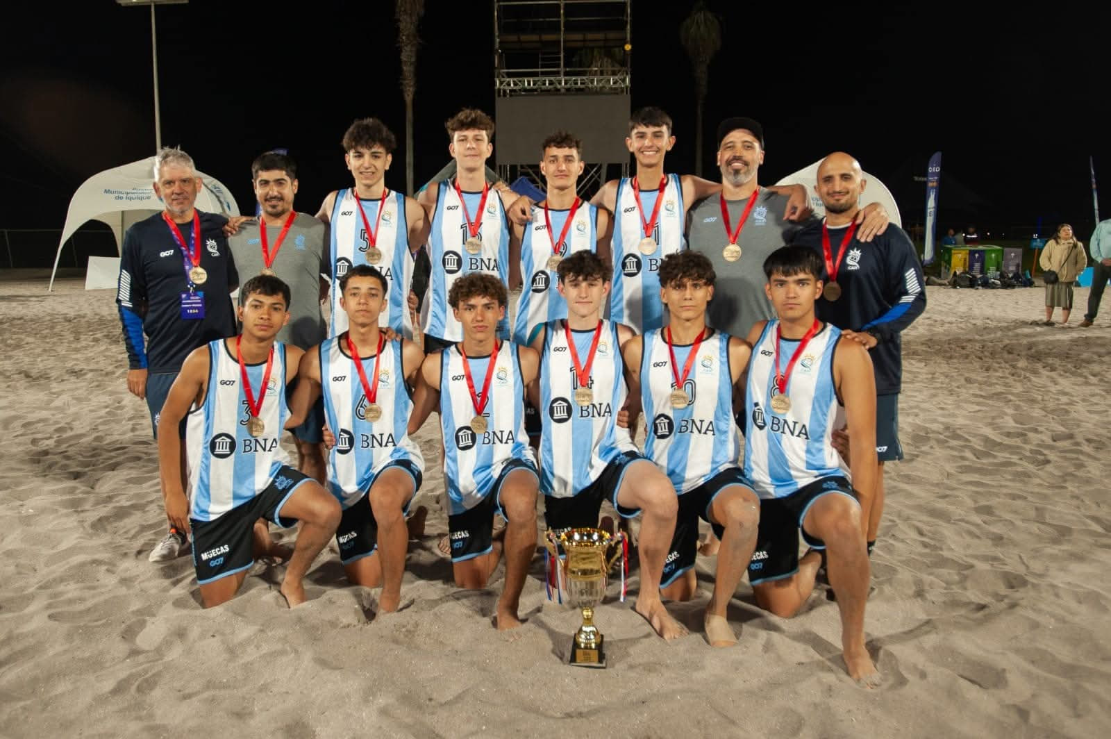 Equipo juvenil de beach handball de Patagones posa en la playa durante la noche con medallas y el trofeo del torneo, junto a sus entrenadores