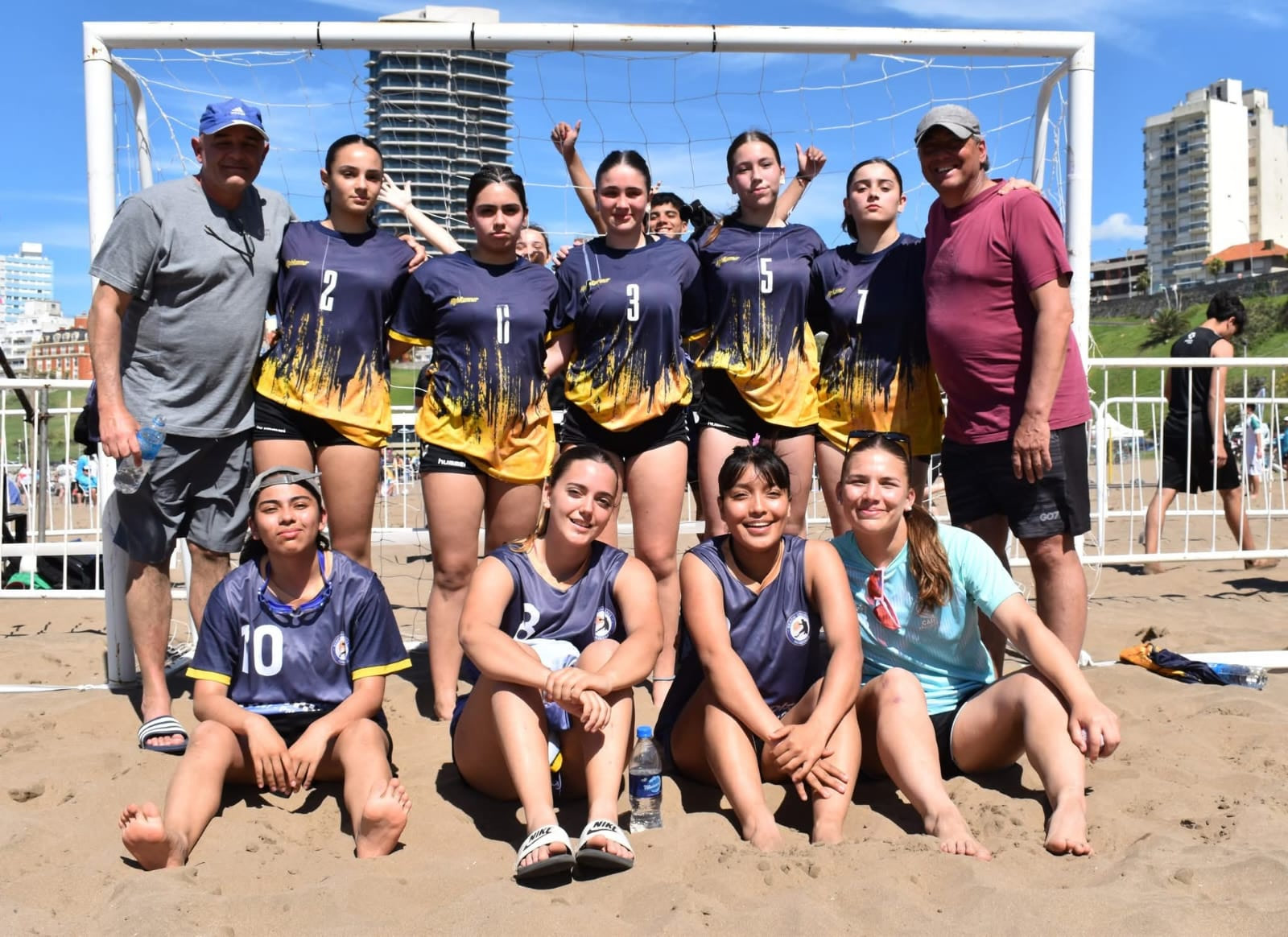 Equipo femenino de beach handball de Patagones posa con sus entrenadores delante del arco, en la arena, con edificios de la costa de fondo
