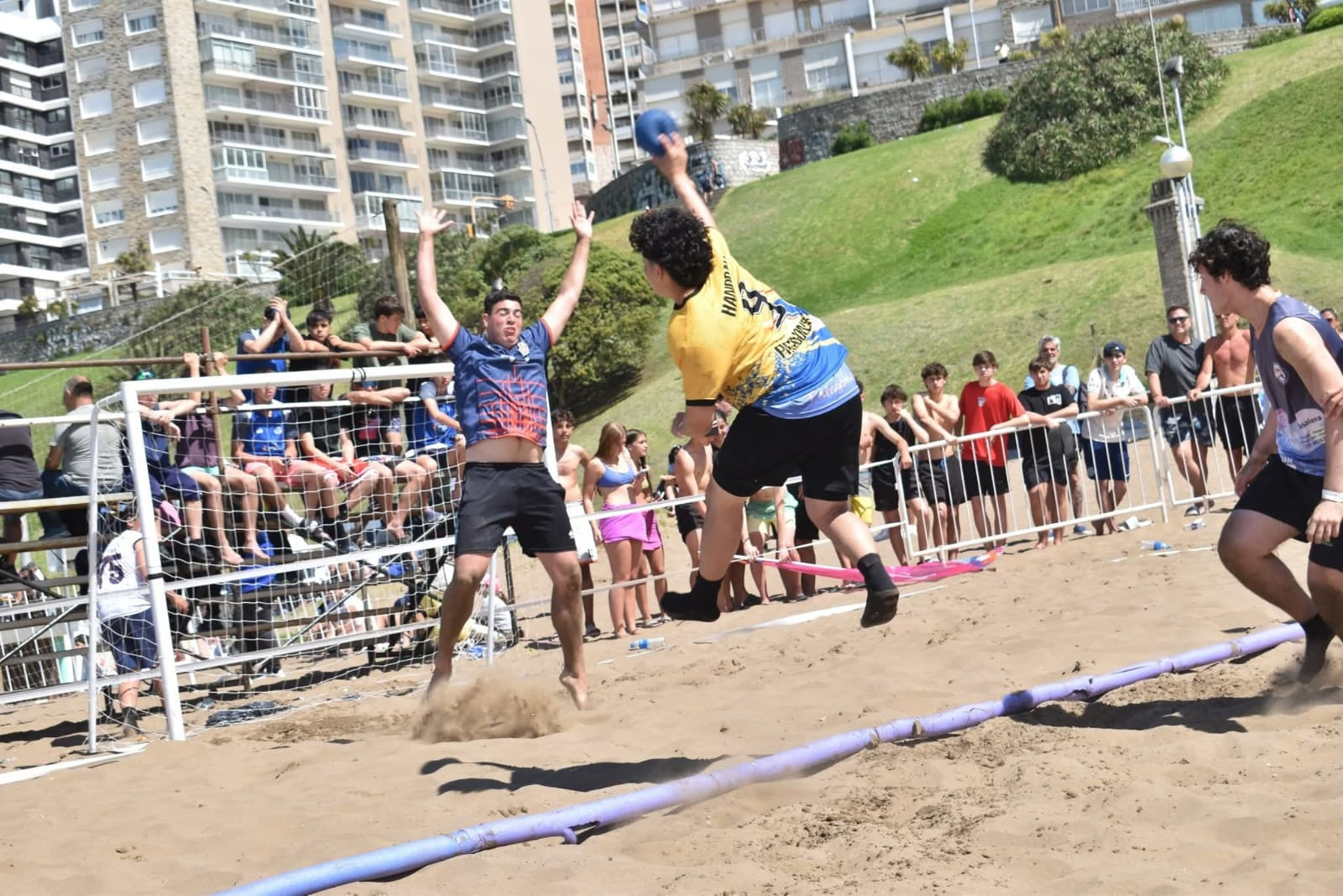Jugador de Patagones con camiseta amarilla salta para lanzar al arco en un partido de beach handball, ante la defensa rival y el público detrás de la reja
