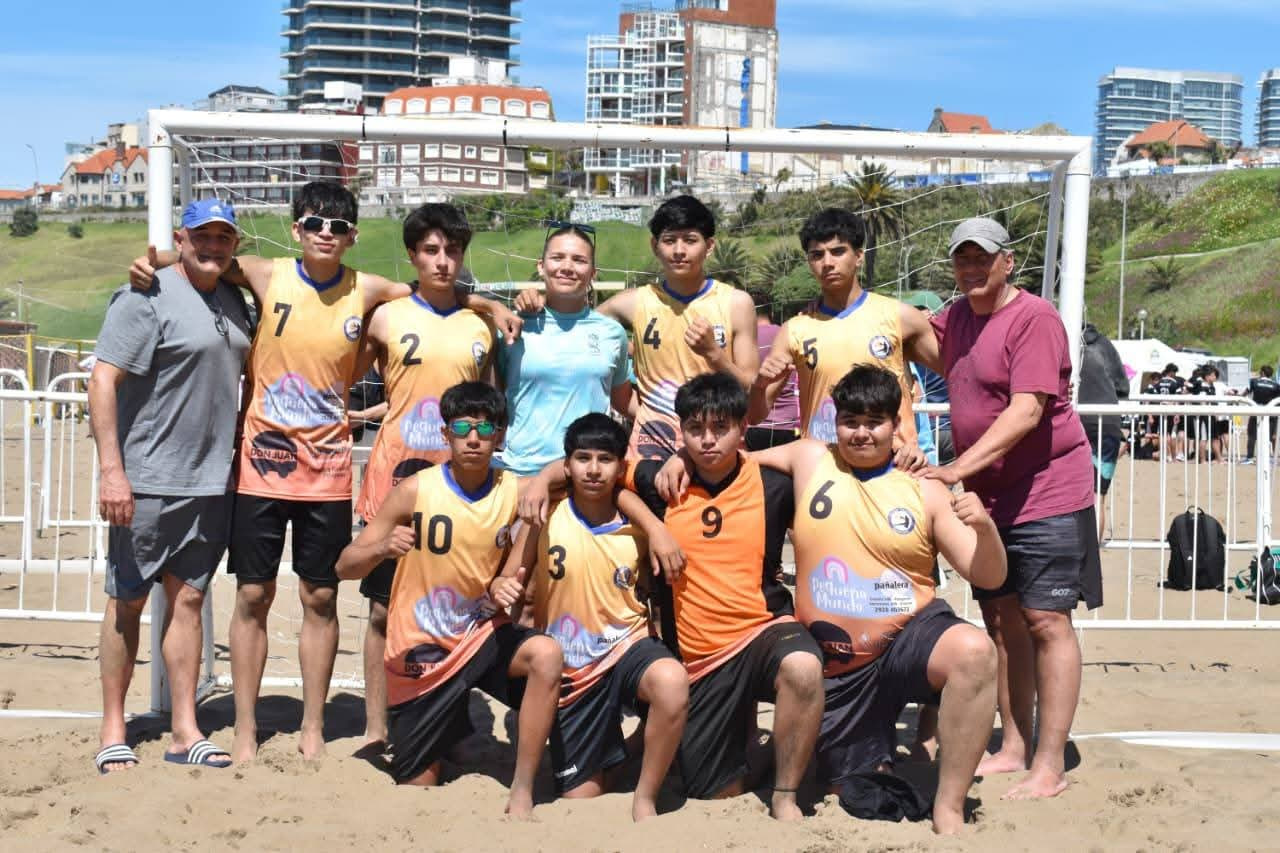 Equipo masculino juvenil de beach handball de Patagones posa con sus entrenadores delante del arco en la playa, con los edificios de la ciudad al fondo