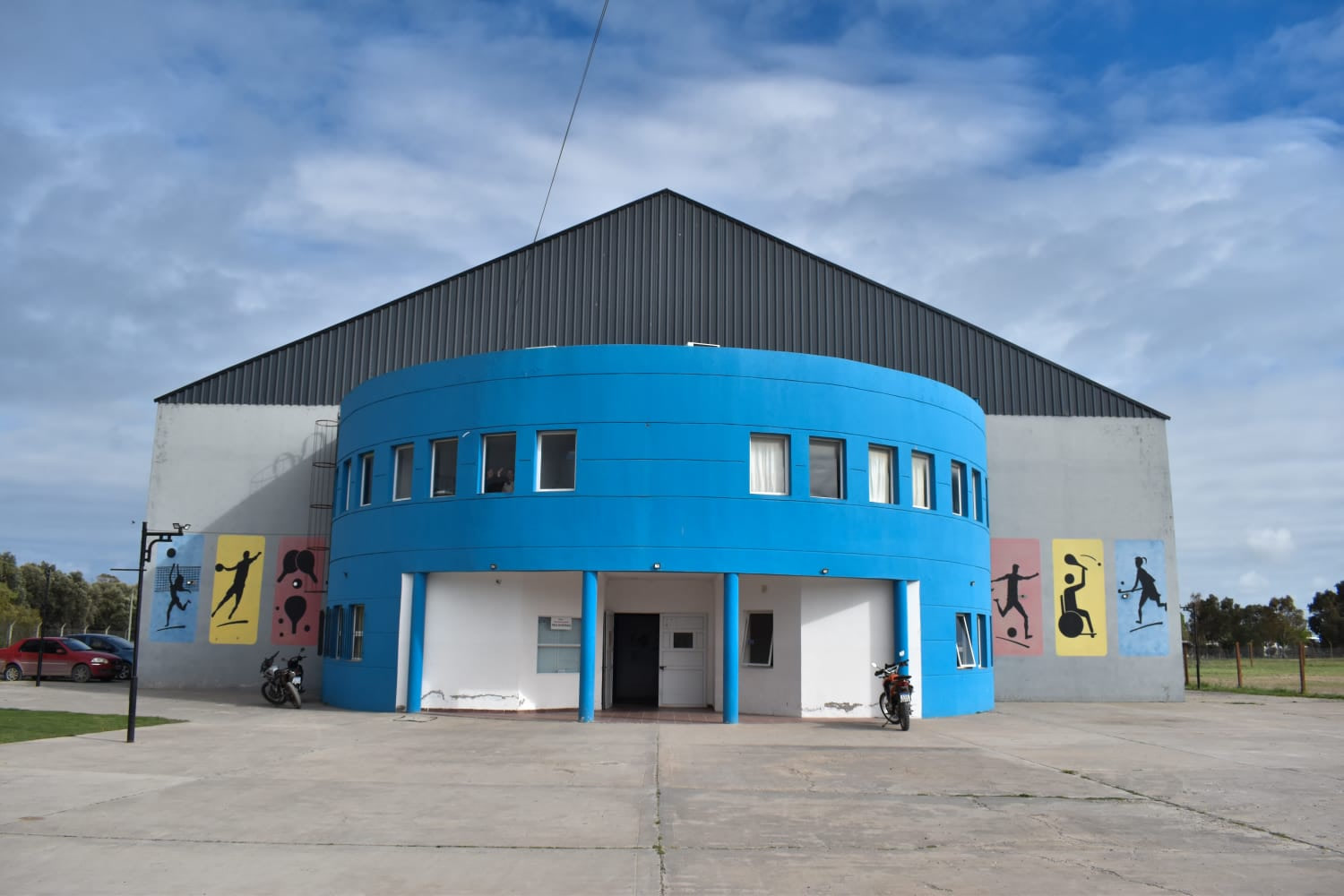 Frente de un edificio deportivo con fachada azul curva y murales con siluetas de deportistas, entre ellos una persona en silla de ruedas, con motos estacionadas en el acceso